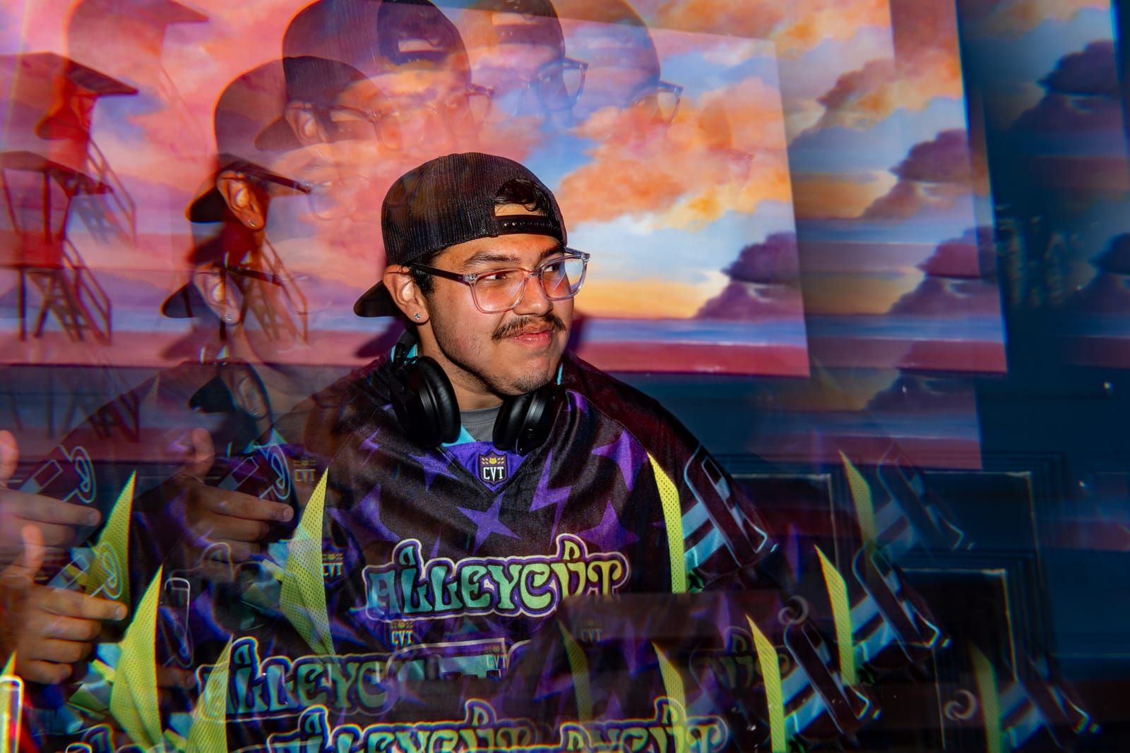 Dill the Menace smiling behind the decks with headphones on, a painted sunset mural behind him