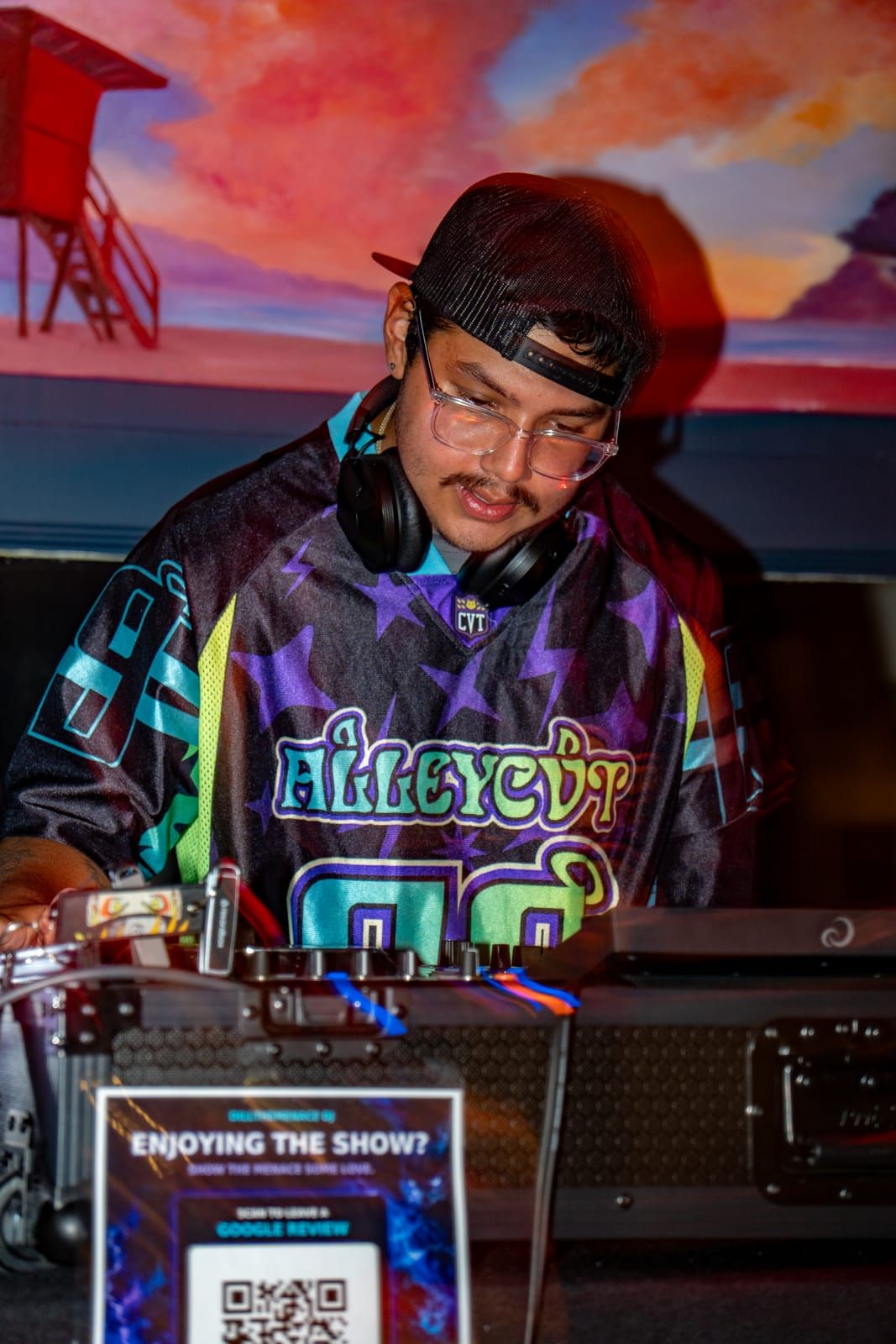 Dill the Menace focused on the mix, headphones on, under the gallery's sunset mural