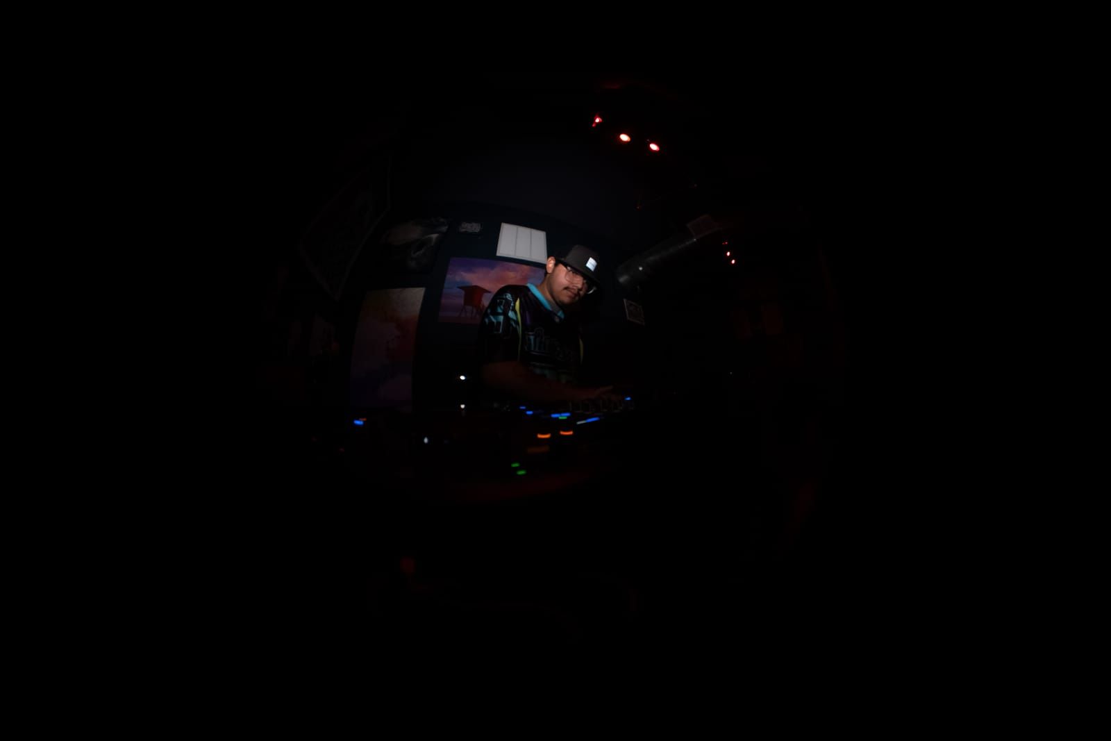 Dimly lit fisheye shot of Dill the Menace behind the decks, controller lights glowing in the dark